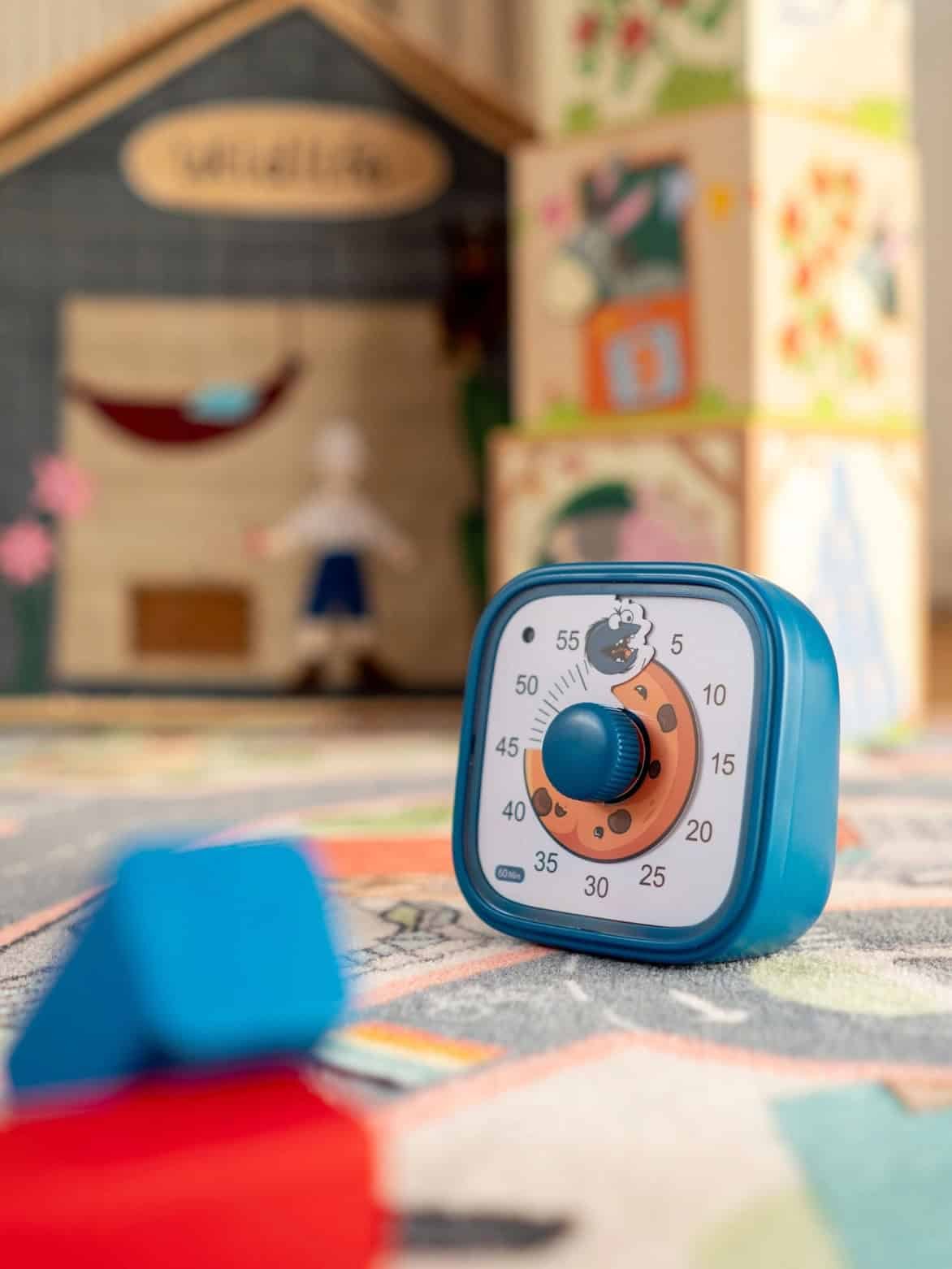 Blue cookie-themed visual timer sitting on a colorful play mat, set to around 38 minutes. In the background, there are large illustrated stacking blocks and a small pretend playhouse. The timer shows a cookie gradually being “eaten” by a cartoon cookie monster character at the top. Let me know if you'd like this in Spanish or need a version optimized for screen readers!